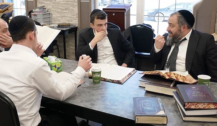 A rav at the head of a table, talking with the young men learning with him, seforim open in front of them.